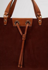 Sac fourre-tout en tissu côtelé marron avec des poignées en cuir beige et un lien en cuir passé à travers des œillets métalliques en haut au centre.