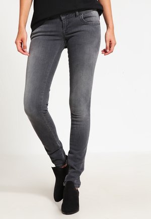 Persoon die strakke grijze spijkerbroek en zwarte enkellaarsjes draagt, met gekruiste benen staand tegen een effen lichte achtergrond.