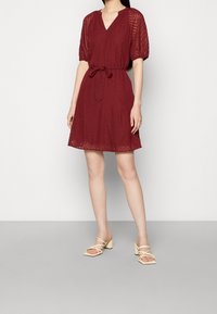 Bourgognefärgad klänning med korta ärmar, v-ringning, elastisk midja och skärp med knytband. Har ett strukturerat tyg med ett subtilt chevronmönster.