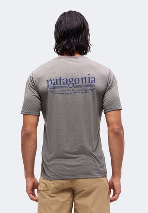 Mand med mørkt mellemlangt hår iført en grå Patagonia t-shirt og beige shorts, vist bagfra mod en ensfarvet lys baggrund.