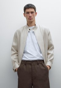 Veste bomber beige avec col montant et fermeture à pression, associée à un t-shirt blanc et un pantalon élastique marron. Texture lisse.