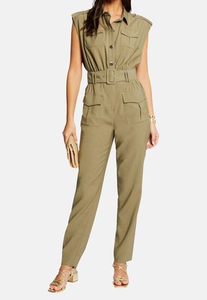 Femme portant une combinaison utilitaire kaki sans manches avec boutons sur le devant, ceinture, plusieurs poches, tenant une pochette tissée et portant des talons dorés à lanières.