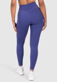 Leggings viola a vita alta realizzati in un tessuto morbido e elasticizzato, con un design delle cuciture sagomato, abbinati a scarpe da ginnastica bianche.