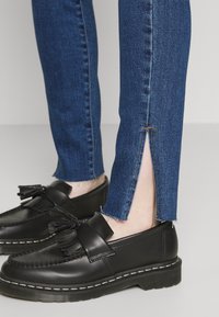 Tmavě modré džíny z denimu s bočním rozparkem na lemu, nošené s černými koženými mokasíny zdobenými střapci a bílým šitím.