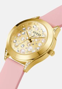Kuldne Guess käekell, millel on pärli ja kristalli aktsendid puutus ja korpus, sobitades pehme roosa silikoonist rihmaga.