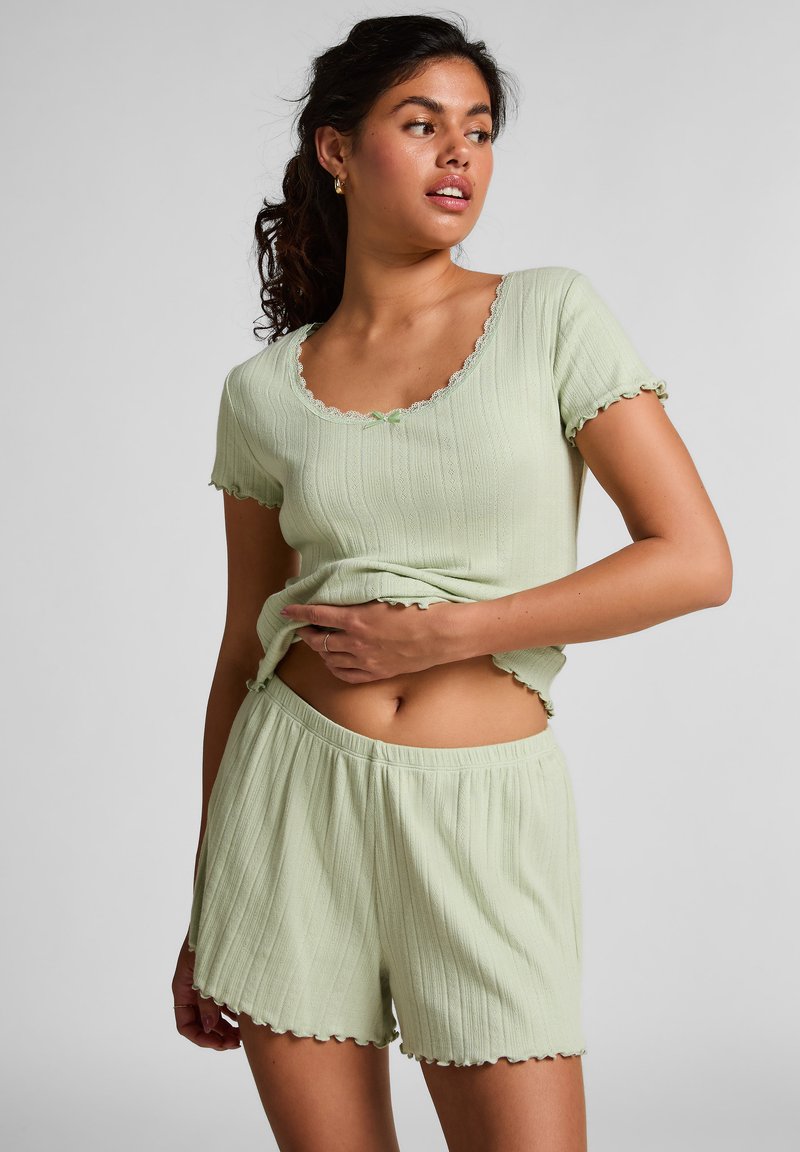 Vrouw draagt een lichtgroene geribbelde top met korte mouwen en bijpassende shorts met een gegolfde rand, met haar hand rustend op haar blootgestelde buik.