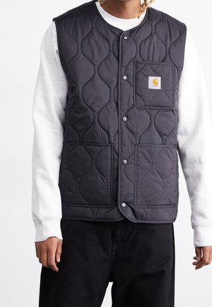 Personne portant une veste matelassée noire avec boutons-pression et une poche avec logo par-dessus une chemise blanche à manches longues et un pantalon noir.