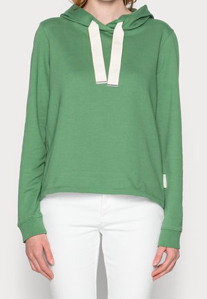 Person trägt einen grünen Kapuzenpullover mit weißen Kordeln und weiße Hosen, steht vor einem schlichten Hintergrund.