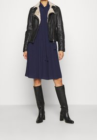 Giacca da moto in pelle nera con colletto in pelliccia sintetica, abbinata a un vestito midi blu navy. Completato con stivali in pelle nera al ginocchio, con un tacco robusto.