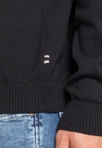 Pull en maille noire avec poignets et ourlet côtelés. Petite étiquette logo avec des rayures rouges, blanches et bleues sur le côté. Porté avec un jean en denim.
