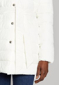 Chaqueta de plumas blanca con cintura ajustada mediante un cinturón, con botones a presión plateados, costuras horizontales y un tejido texturizado.