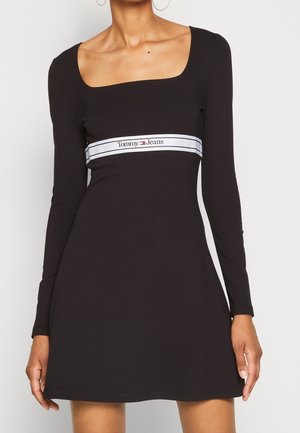 Robe longue noire à manches longues avec un décolleté carré et un corsage ajusté. Elle présente une ceinture blanche de marque avec le logo "Tommy Jeans".