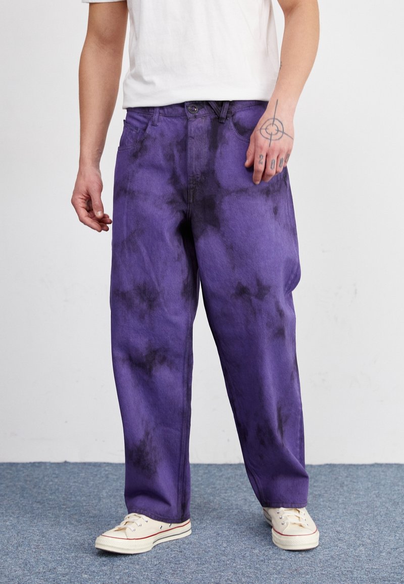 Paarse spijkerbroek met een tie-dye patroon, met een relaxte pasvorm en rechte benen, gecombineerd met witte sneakers en een effen shirt.