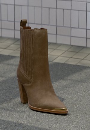 Světle hnědá semišová kotníková bota na podpatku s elastickými bočními panely a kovovým zlatým špičkou, na texturovaném venkovním povrchu.