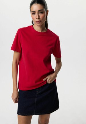 Młoda kobieta z ciemnymi włosami, ubrana w czerwoną koszulkę i ciemną spódnicę dżinsową, stoi z jedną ręką w kieszeni spódnicy na jednolitym tle.