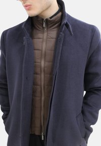 Cappotto di lana navy sovrapposto a una giacca imbottita marrone con zip, dotata di colletto rialzato e tasche laterali. Il tessuto appare morbido con una superficie liscia.