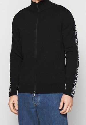 Hombre con jersey negro con cremallera y texto "KARL LAGERFELD" en las mangas, combinado con vaqueros azules, de pie frente a un fondo liso.
