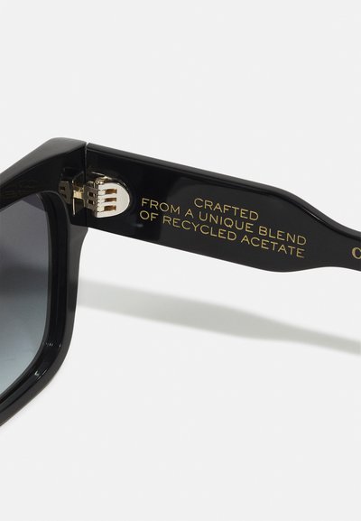 Black sunglasses with a rectangular frame made from recycled acetate. Features include a gold accent on the arm and gradient dark lenses.