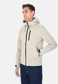 Giacca beige chiaro con cappuccio, due tasche frontali con zip, dettagli neri e un design elegante e moderno. Presenta un tessuto liscio e testurizzato.