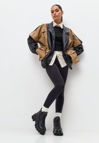 Bruine en zwarte oversized jas met drukknoopsluiting over een zwarte top, wit- en zwartgeruite shirt, zwarte leggings en chunky zwarte laarzen.