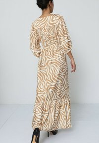 Robe maxi beige avec des rayures blanches en zèbre, présentant des manches bouffantes, une taille élastique et un ourlet volanté. Des talons noirs sont portés.