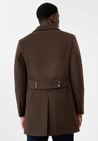 Cappotto in lana marrone con un design su misura, cintura sul retro con bottoni decorativi e spalle strutturate. Texture liscia con una finitura ordinata.