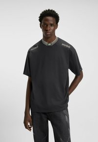 Mørkegrå kortærmet T-shirt med ribbet krave, prydet med lys grå "HUGO" branding på skuldrene. Glat stofstruktur.