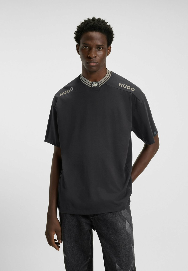 Mørkegrå kortærmet T-shirt med ribbet krave, prydet med lys grå "HUGO" branding på skuldrene. Glat stofstruktur.
