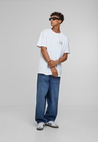 Hvid kortærmet t-shirt med logo, parret med løstsiddende blå denim jeans og grå sneakers med sort og hvid striber. Sorte solbriller.