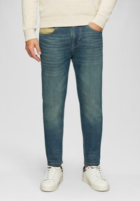 Jeans en denim bleu clair avec une coupe slim, mettant en avant une poche jaune, portés avec des baskets blanches et un pull crème.