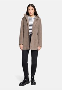 Bruine fleece jas met een capuchon, voorzakken, gecombineerd met een gestreept shirt en zwarte jeans, gedragen met zwarte enkellaarsjes.