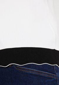 Suéter blanco con canalé y dobladillo negro con forma de ondas, tejido texturizado, combinado con vaqueros con costuras naranjas.