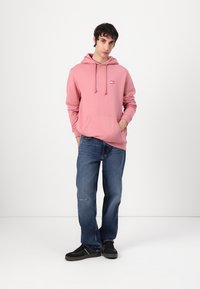 Felpa rosa con tasca a marsupio e cappuccio regolabile, abbinata a jeans blu e scarpe nere. Texture morbida, vestibilità casual.