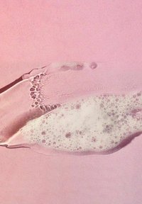 Lyserød fløde med hvide bobler, glat tekstur og en blød glans, påført over en lys pink baggrund, der skaber subtile mønstre.