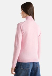 Femme aux longs cheveux bruns portant un pull à col roulé rose clair et un jean bleu, de dos contre un fond blanc.
