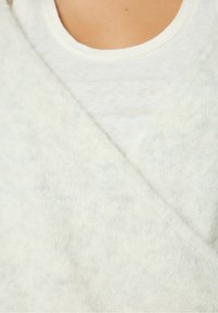 Weißer, weicher, strukturierter Überwurf mit V-Ausschnitt, der über einem schlichten weißen Shirt getragen wird. Insgesamt helle, neutrale Farbpalette.