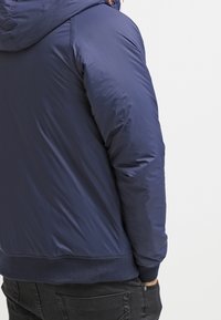 Giacca a vento trapuntata blu navy con cappuccio, caratterizzata da un esterno liscio e lucido, polsini a costine e fascia elastica in vita.