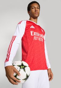 Rød og hvid langærmet fodboldtrøje med tekstureret mønster, med "Emirates" logo. Holder en sort og hvid fodbold med stjernemønster.