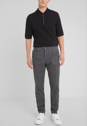 Hombre con polo negro de manga corta con cremallera, pantalones grises plisados y zapatillas blancas, de pie frente a un fondo liso gris claro.