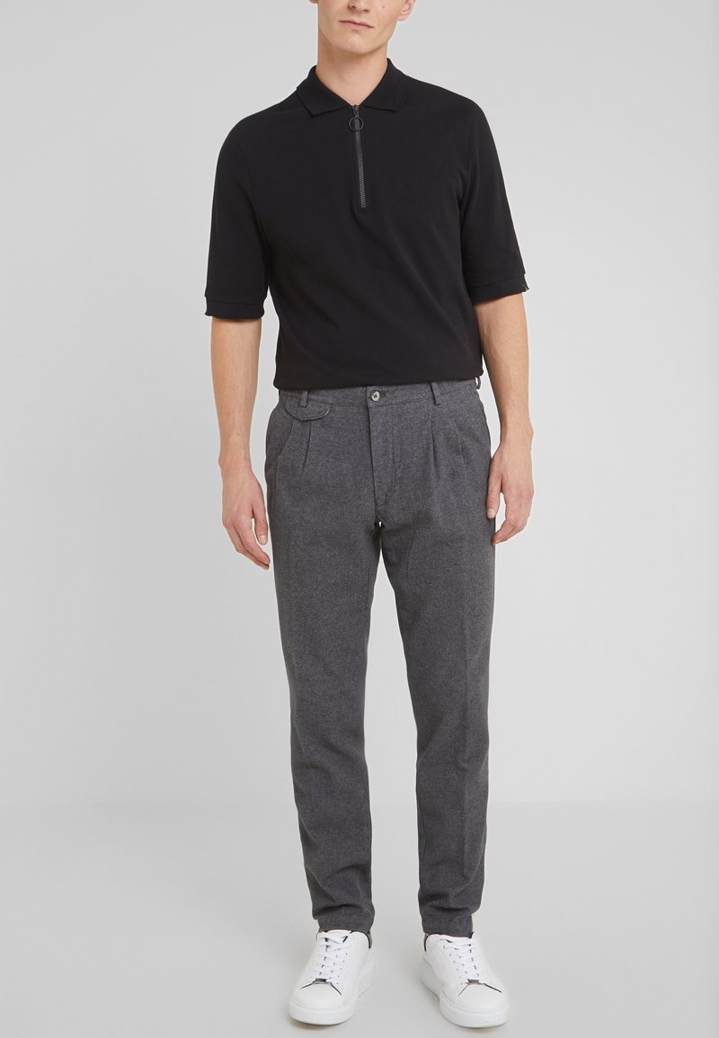 Homme portant un polo noir à manches courtes avec fermeture éclair, pantalon gris plissé et baskets blanches, debout devant un fond gris clair uni.