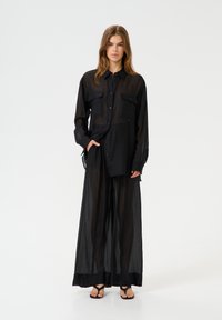 Black sheer shirt with two front pockets, collar, and button closure, paired with matching loose-fitting pants and flat sandals.