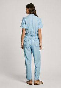 Light blue denim jumpsuit with short sleeves, elastic waist, and two back pockets. Features a collar and relaxed fit. Flat sandals worn.