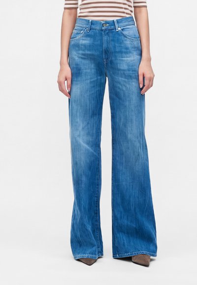 Femme portant un jean large taille haute bleu clair avec poches sur le devant et fermeture par boutons, associé à des chaussures marron pointues et un haut rayé.