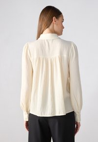 Blusa de crema clara, con textura, y cuello abotonado, con fruncidos en la parte trasera, y mangas largas con puños abotonados. Combinada con pantalones oscuros.
