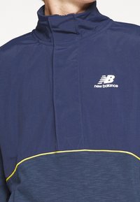 Tmavě modrý pulovr s půlvýstřihem na zip, vyrobený z lehkého materiálu, s žlutým akcentem podél spodního okraje a bílým logem New Balance na hrudi.