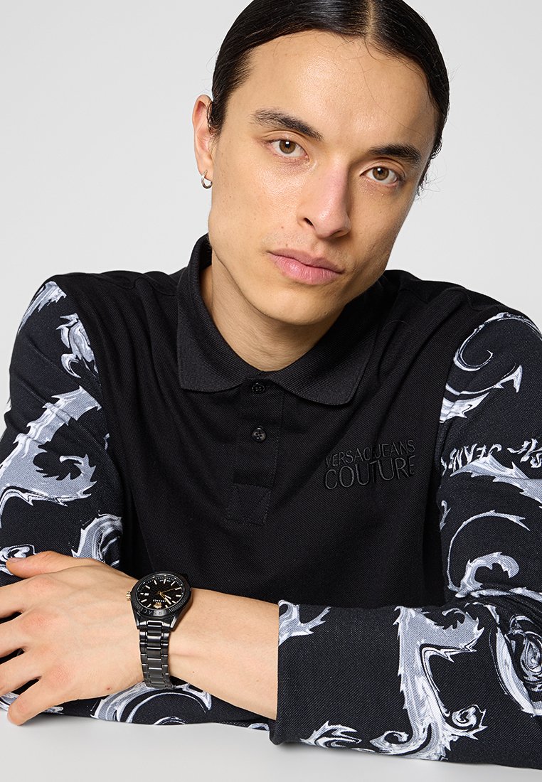 Schwarzes Polohemd mit kontrastierenden, gemusterten langen Ärmeln, gefertigt aus einem strukturierten Stoff und mit gesticktem Logo. Eine schwarze Armbanduhr rundet den Look ab.