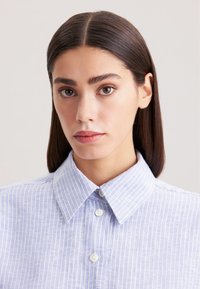 Camicia col celeste chiaro con strisce bianche verticali, chiusura frontale con bottoni, texture liscia e design del colletto standard, focalizzandosi su un aspetto curato.