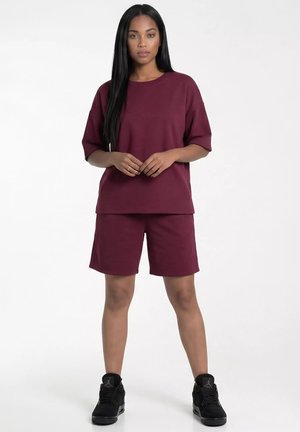 Kobieta stojąca w dopasowanej bordowej oversize koszulce i szortach sięgających kolan oraz czarnych wysokich trampkach.