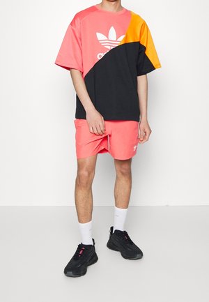Mies, joka pitää väriblokkipainatuksella varustettua Adidas-paitaa väreissä koralli, musta ja oranssi, korallinväriset shortsit, valkoiset sukat ja mustat lenkkarit valkoista taustaa vasten.