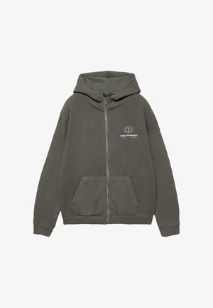 Temno zelena jopa z zadrgo, sprednjimi žepi in belim napisom "Project Division Dept." ter logotipom na levi strani prsnega koša.